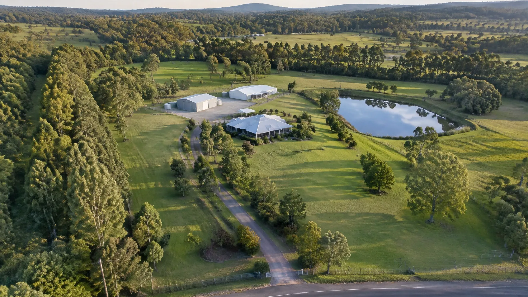 Golden-hour aerial view of an acreage property with a home, sheds, dam, driveway and green rural land